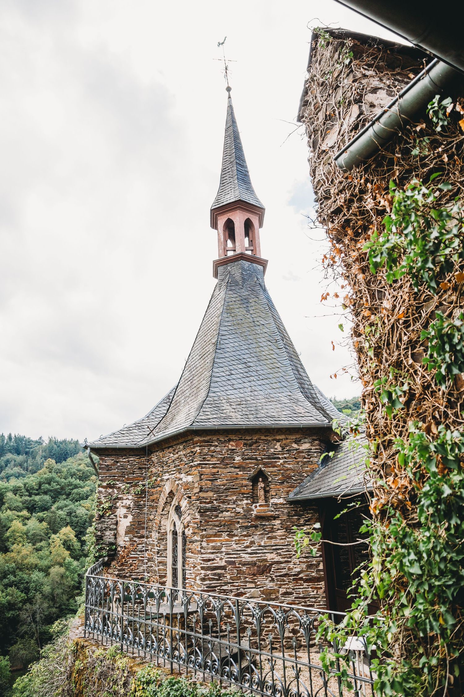Gothic stone chapel with pointed spire and slate roof perched on a hillside covered in ivy.