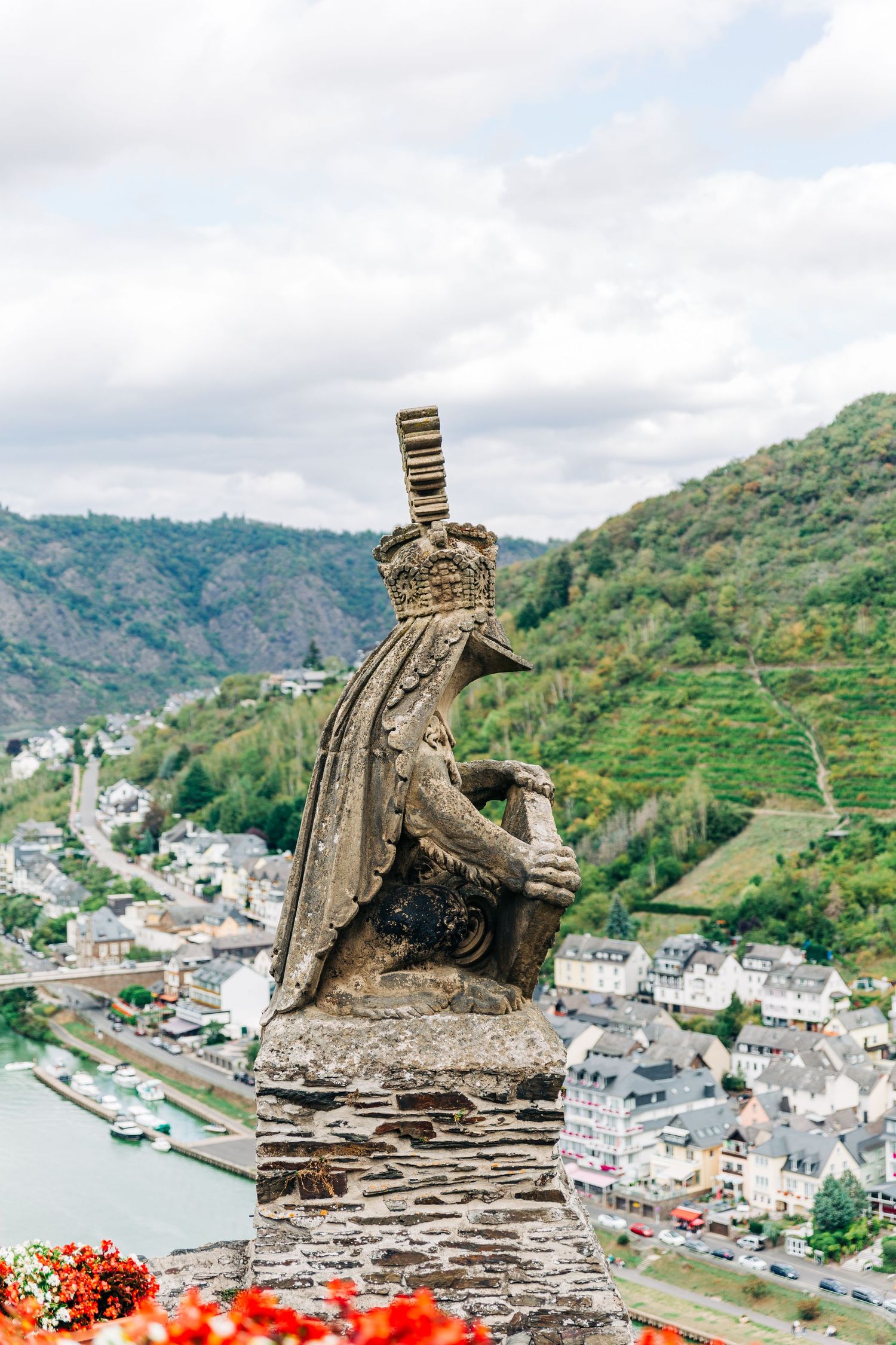 Stone statue of a crowned figure overlooking river valley with terraced hillsides and town below.