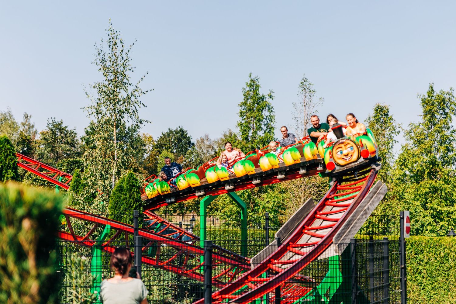 Colorful children's roller coaster train climbing track surrounded by green trees.
