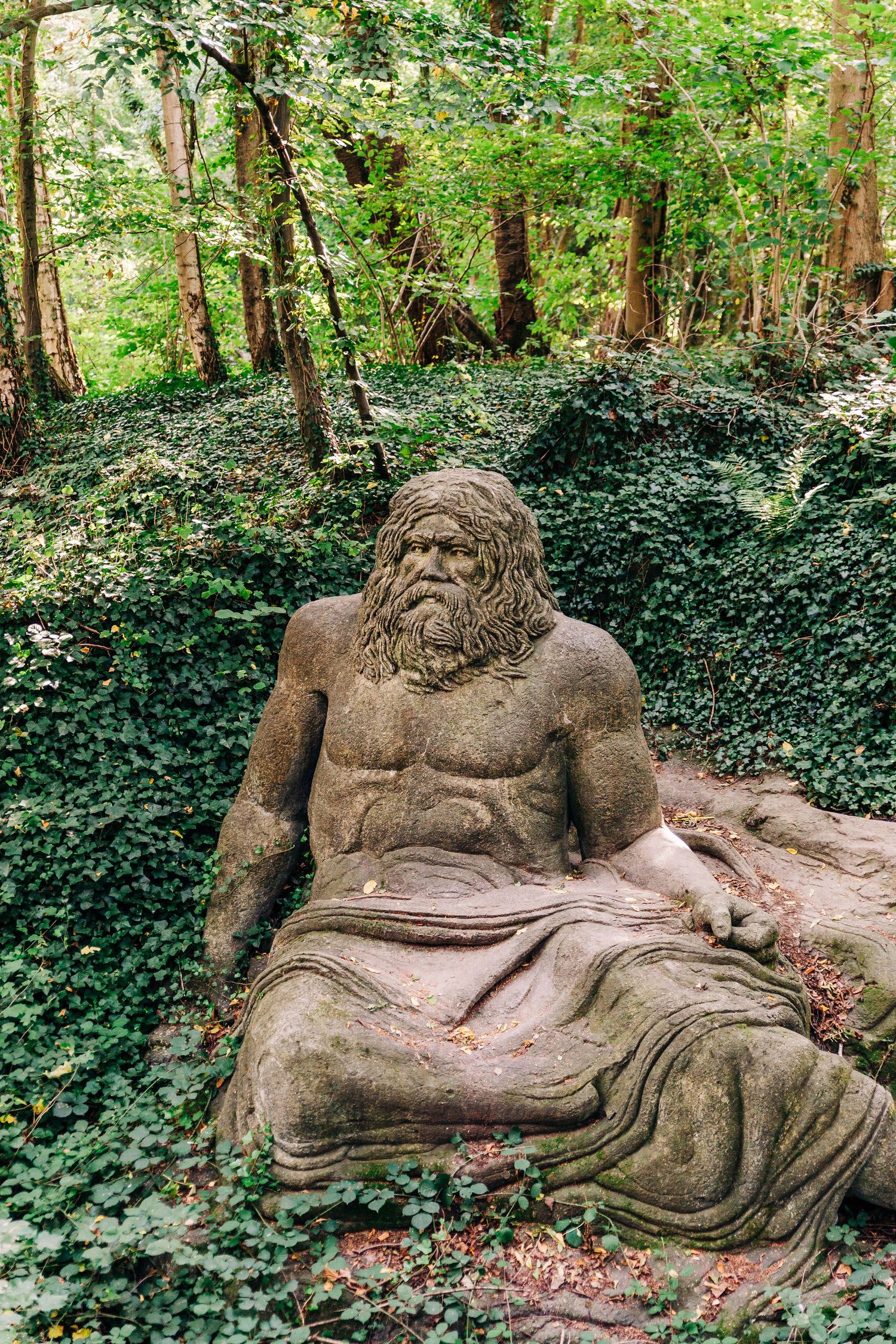 Stone sculpture of a bearded figure reclining in an ivy-covered woodland setting.
