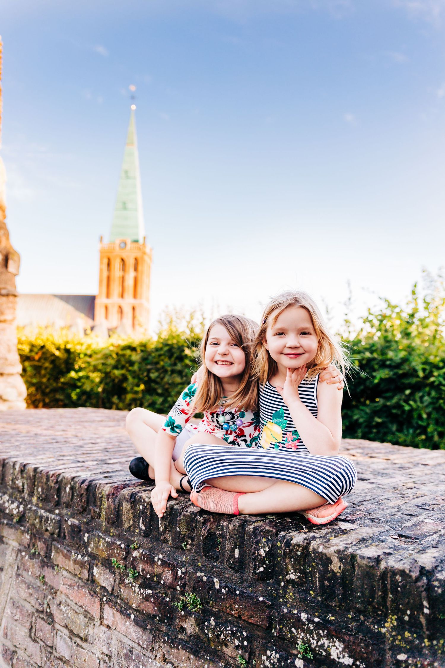 Two children sitting on an old stone wall with a church spire visible in the background.