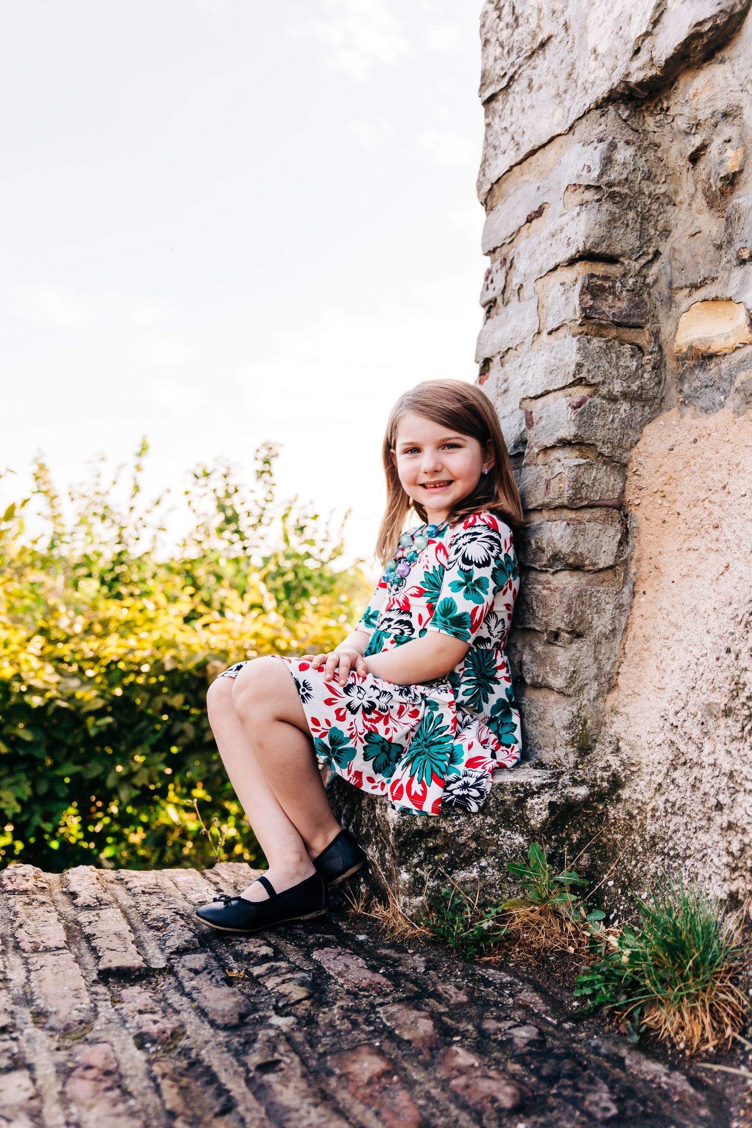 Child in a floral dress sitting on a stone wall during golden hour.