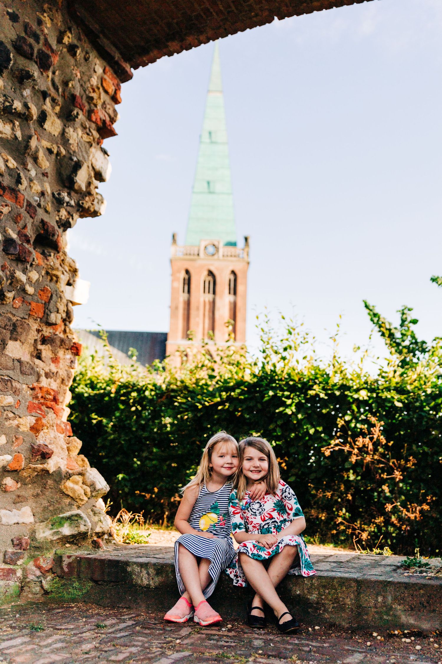 Children sitting together on old stone wall with church spire visible through archway.
