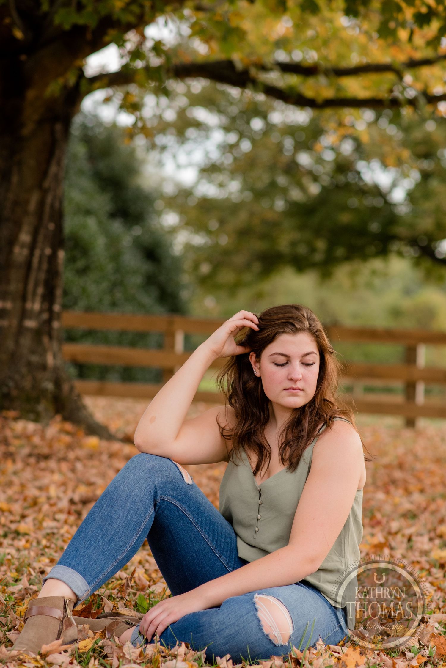 Woman in casual attire sitting on fallen autumn leaves beneath a tree in afternoon sunlight.