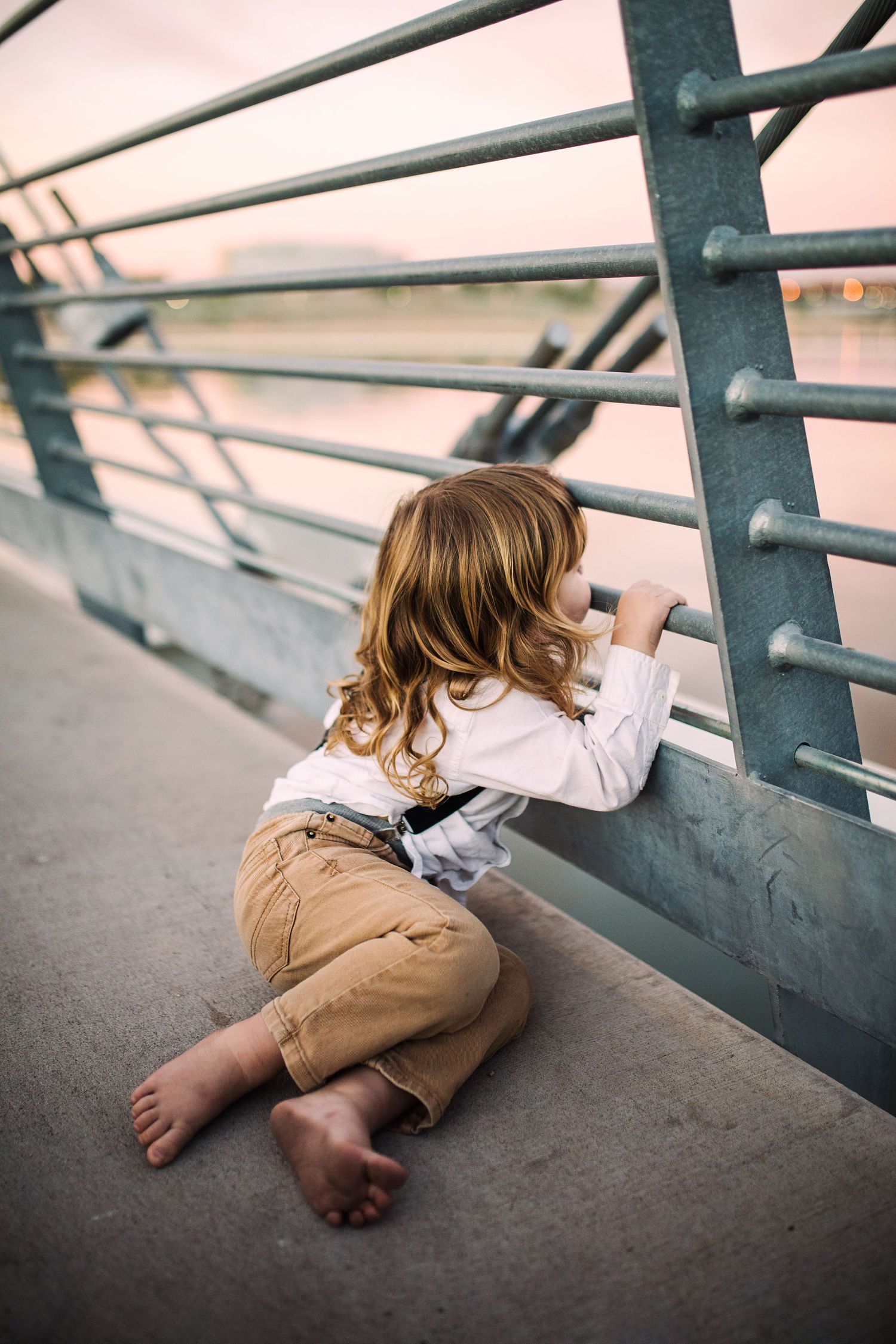 Barefoot toddler with curly hair sitting on bridge looking through metal railing at water below.