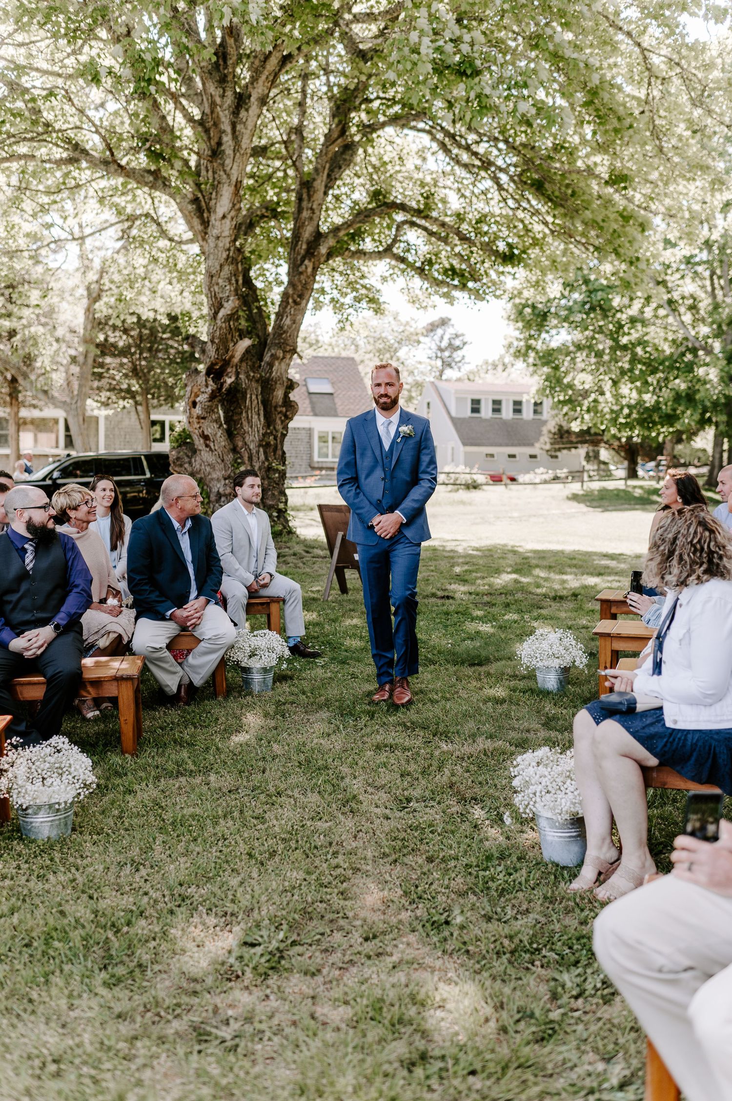 Wedding guests sit on wooden benches under shady trees during an outdoor ceremony.