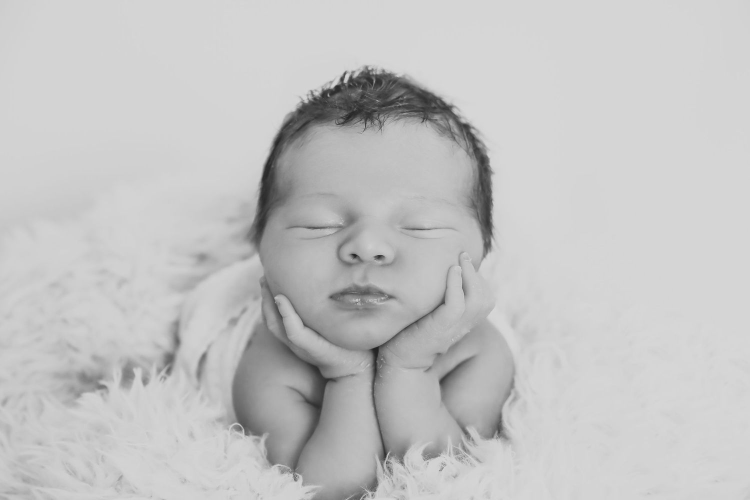 A peaceful newborn baby resting on white fur with hands gently cupping their face.