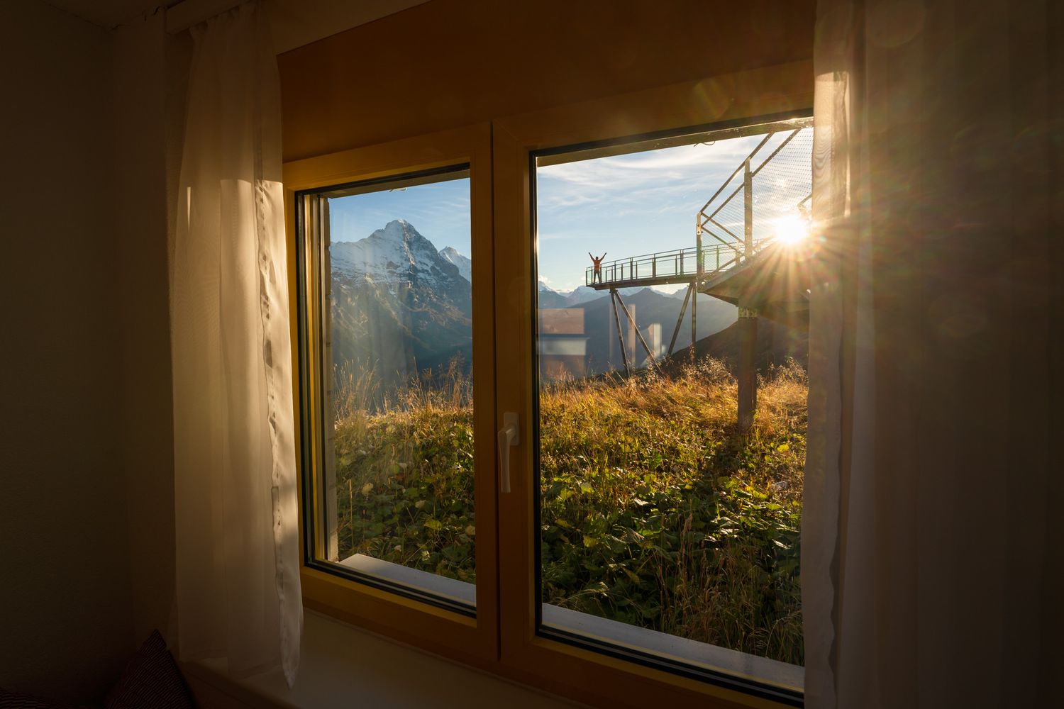 Golden sunlight streams through a window overlooking mountain peaks and wildflowers at sunset.