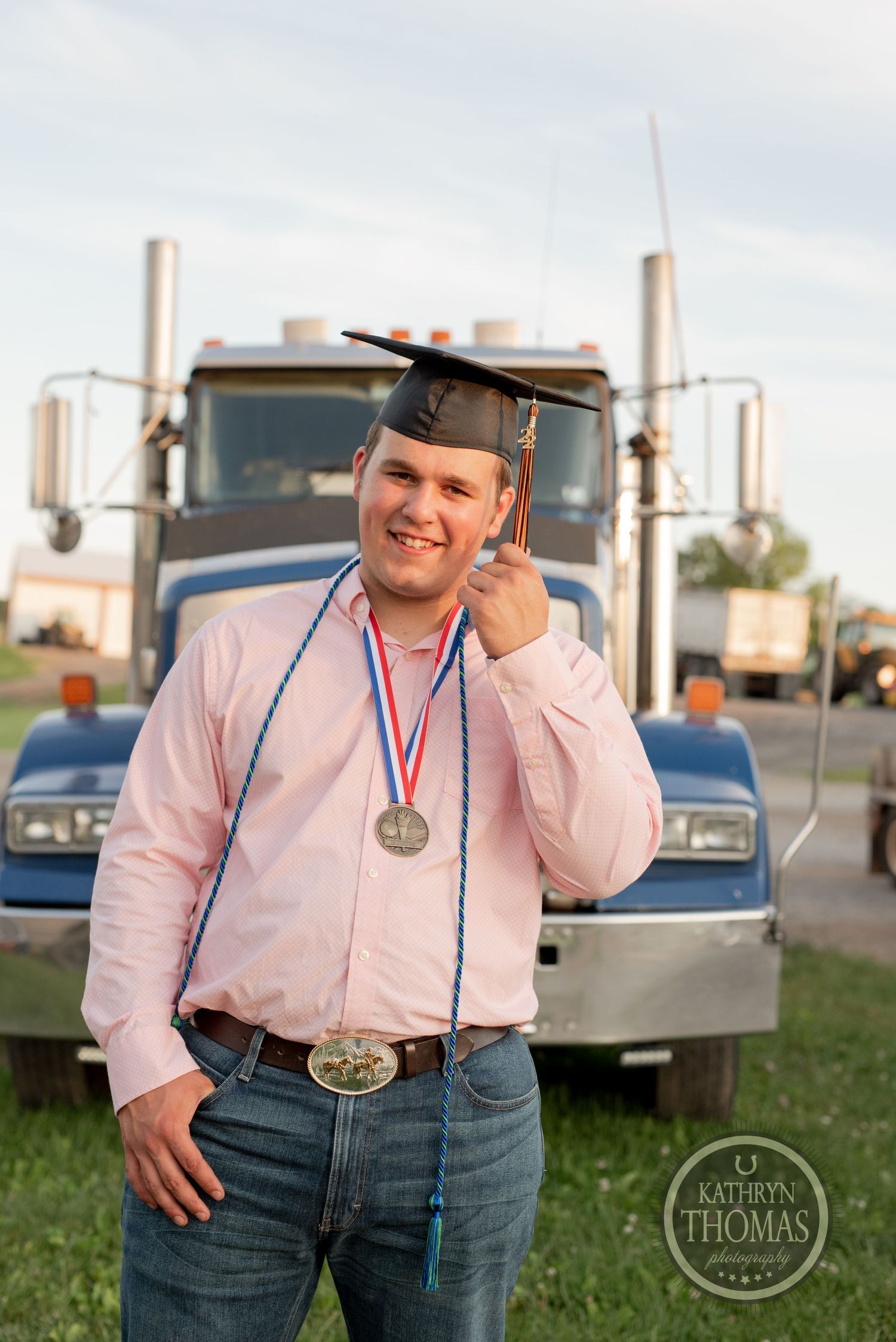 Graduate in pink shirt and cap posing with medal in front of semi truck.