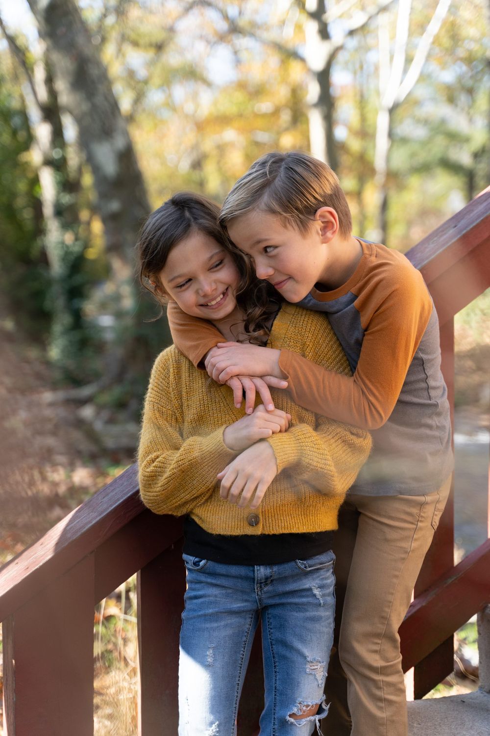 Two young friends share a playful embrace on a wooden bridge surrounded by autumn trees.