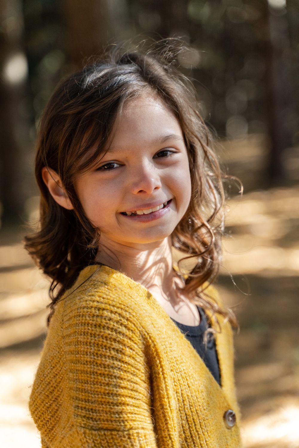 Young person in yellow cardigan smiling warmly in dappled forest sunlight.