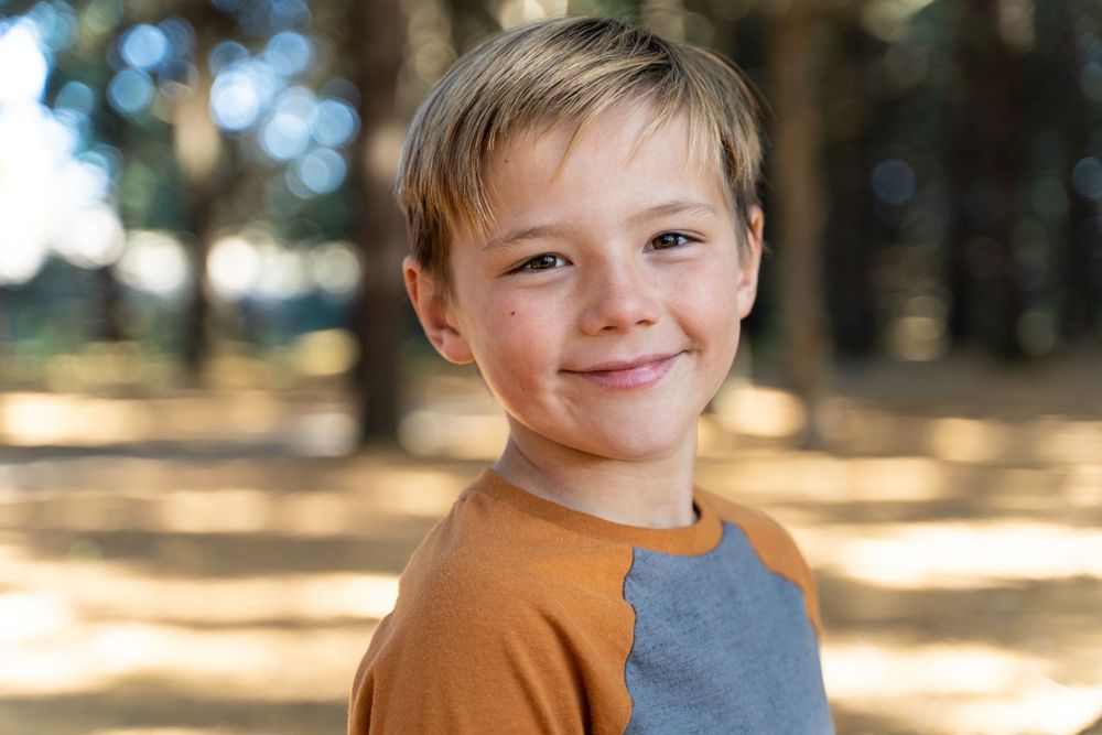 Young person in orange and grey shirt smiles cheerfully in sunny forest setting.