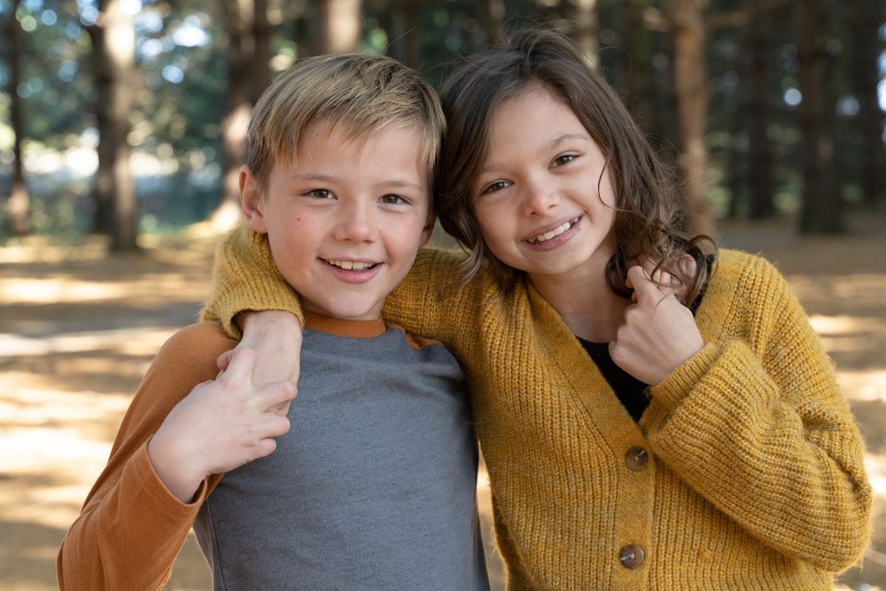Two friends pose together in a forest, wearing casual autumn clothing.