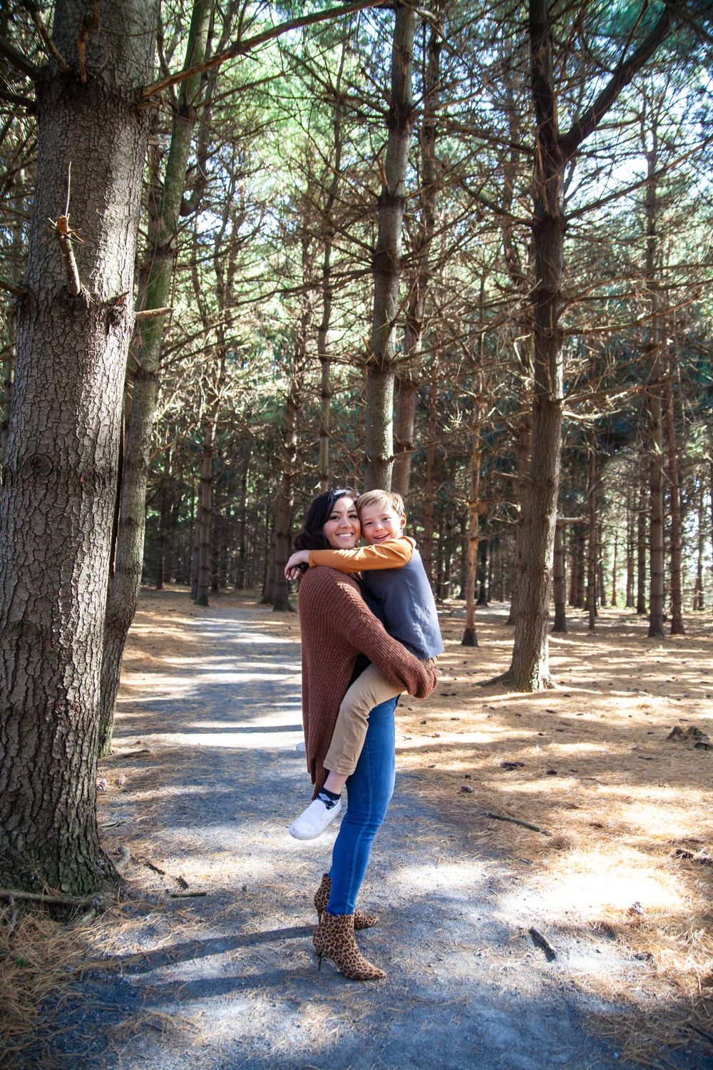 Two people share a joyful embrace on a sunlit forest path surrounded by pine trees.