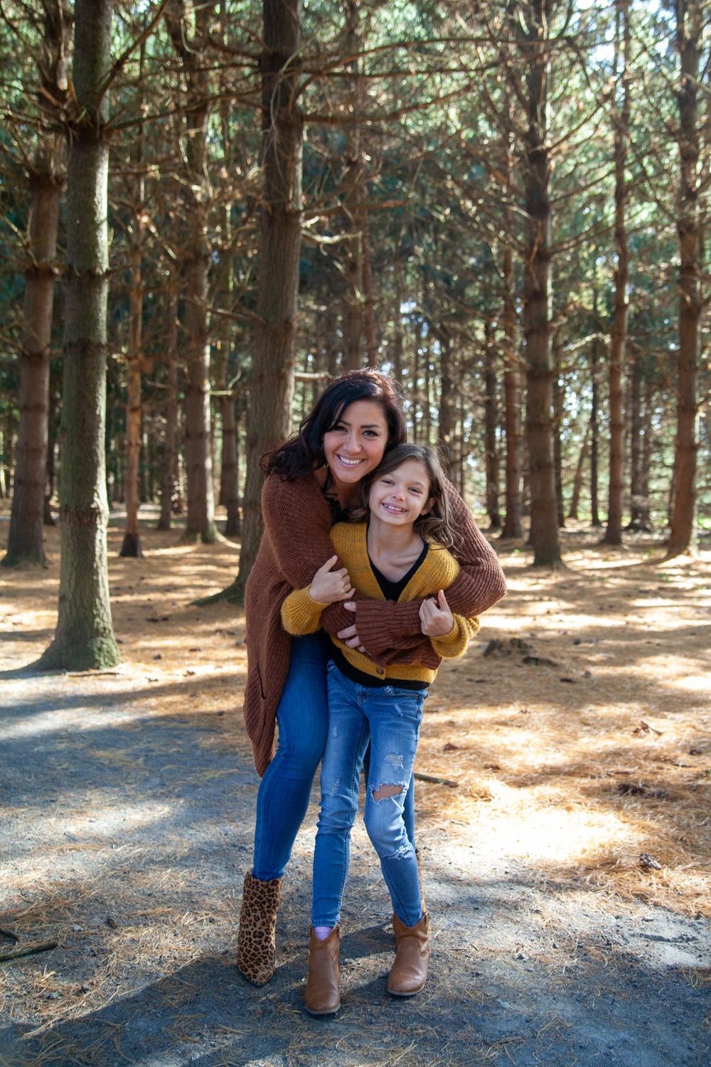 Two people sharing a warm hug while standing on a forest path surrounded by tall pine trees.