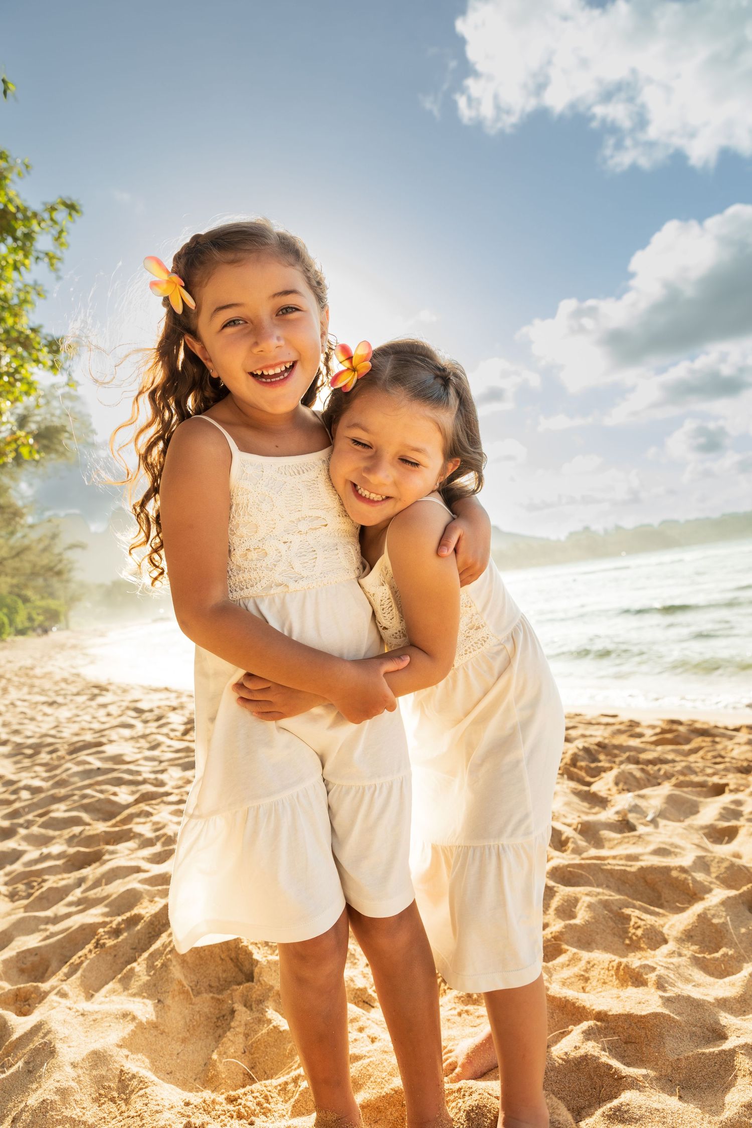 Two girls in white dresses share a joyful embrace on a sunny beach.