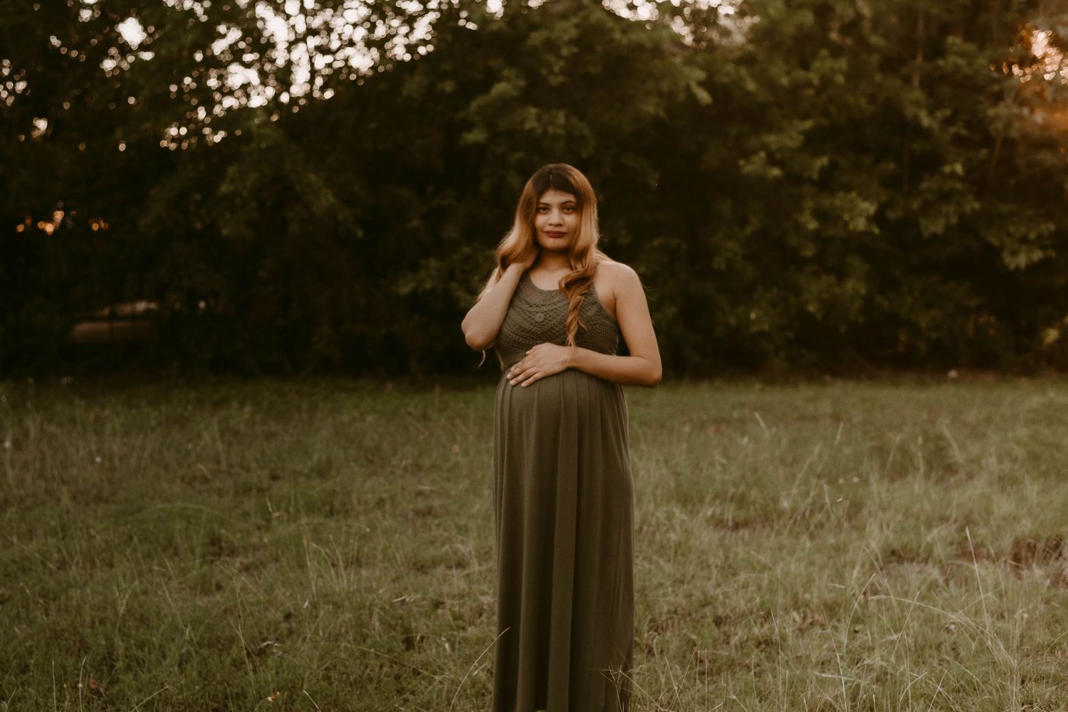 Maternity portrait in olive maxi dress against sunset-lit trees and grass.