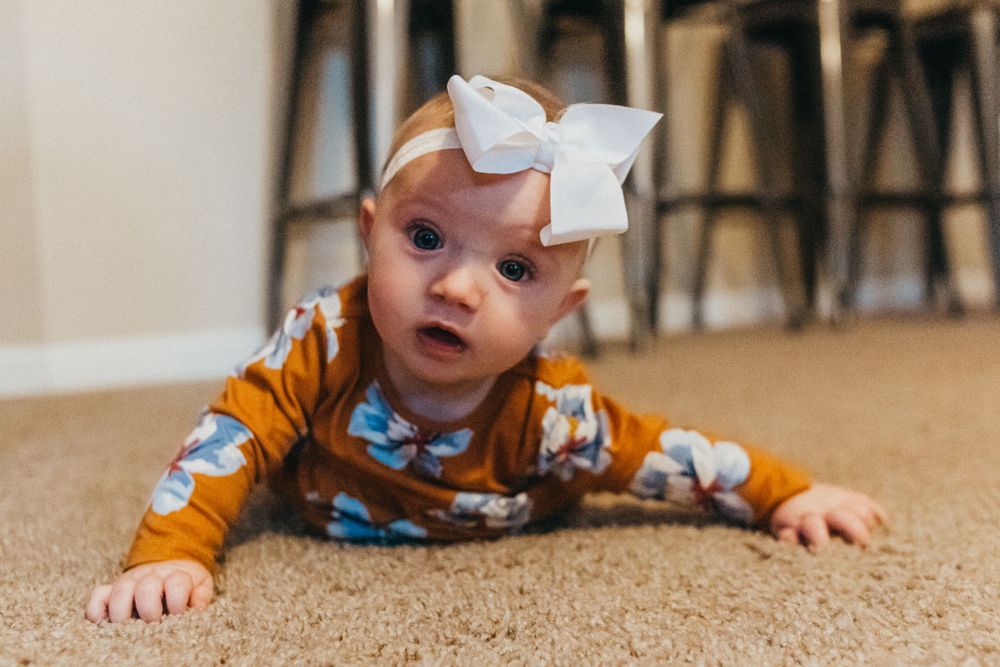 Baby with white bow crawling on beige carpet wearing orange floral outfit.