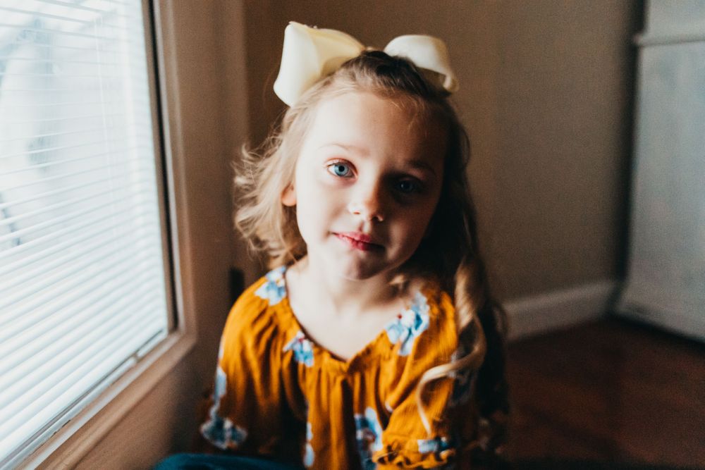 A child in a mustard yellow floral dress with a white bow sits by a window.