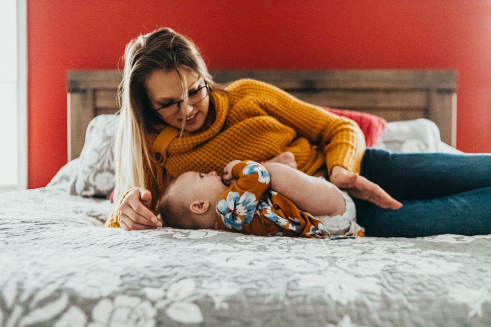 Parent and baby share playful moment together on a cozy bed with white bedding and red wall backdrop.
