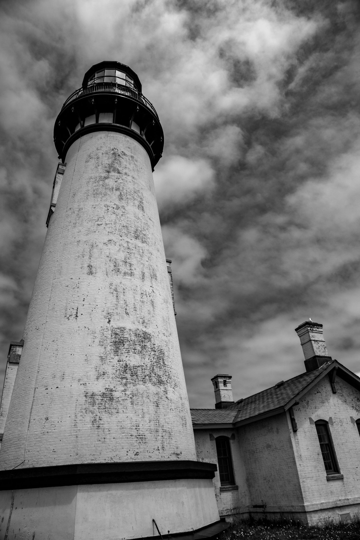 A tall white lighthouse tower stands against dramatic cloudy skies in black and white.