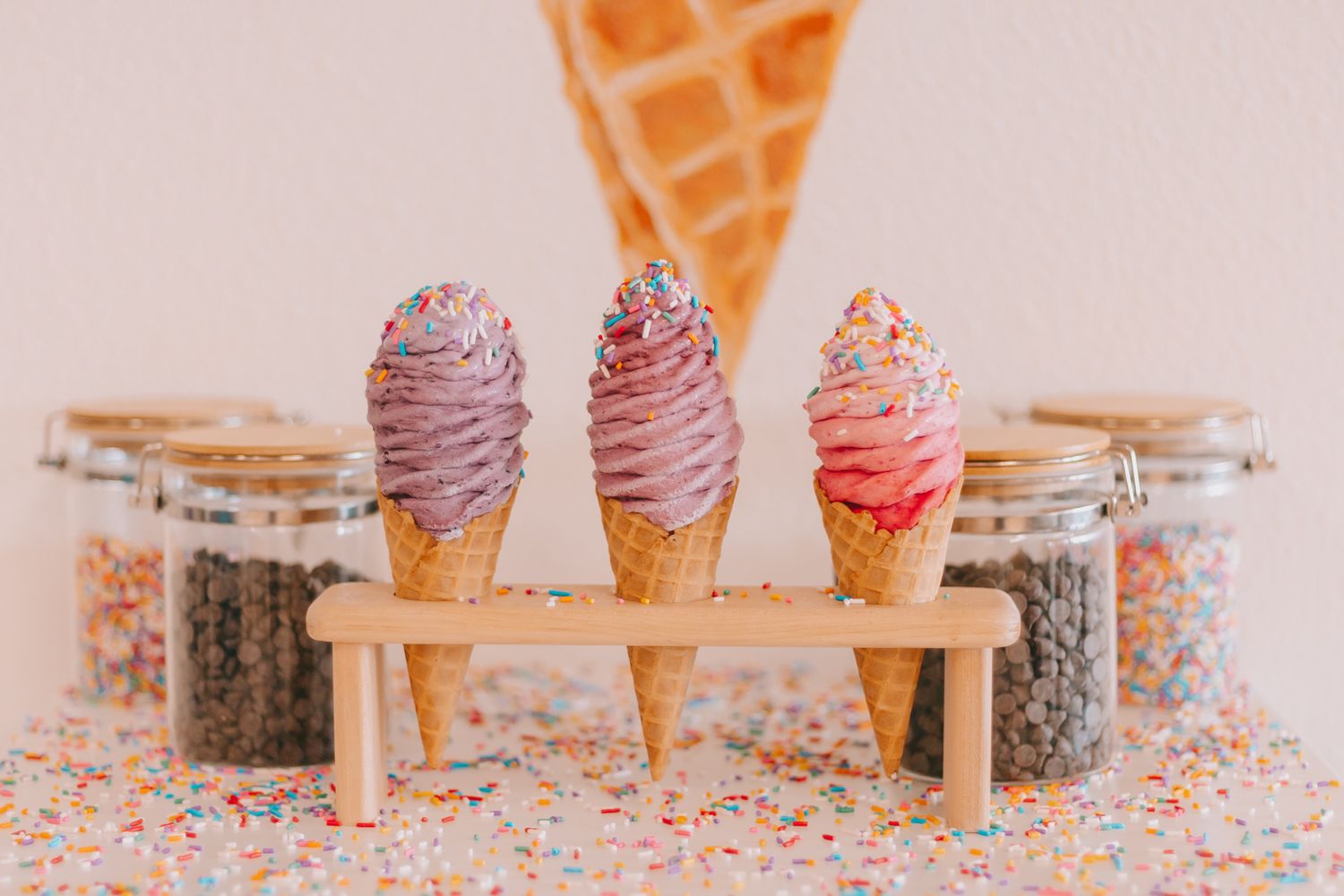 A tray of 3 ice cream cones with jars of toppings in background on a a table covered in sprinkles.