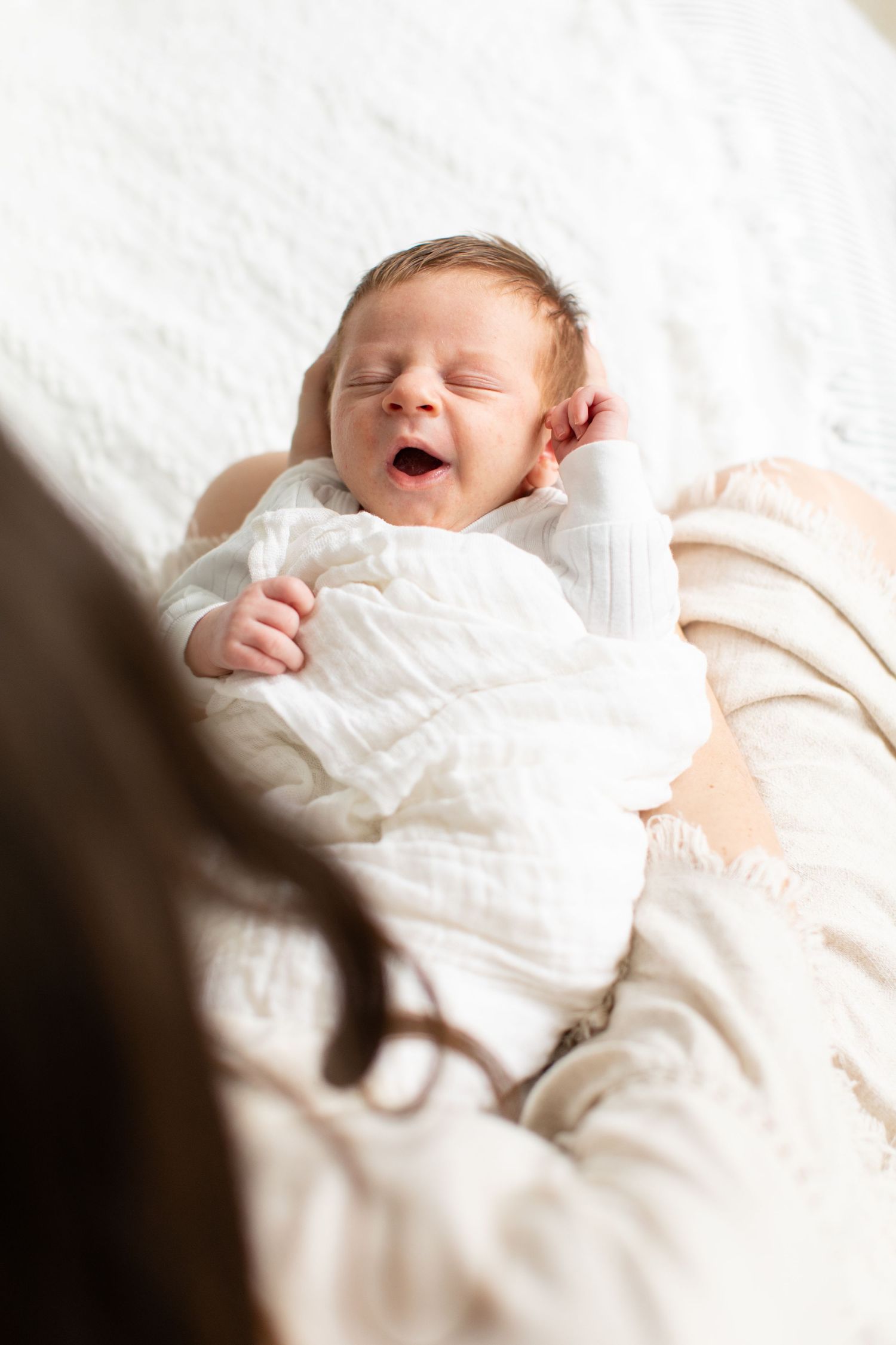 Newborn baby yawning while wrapped in white swaddle on light bedding.