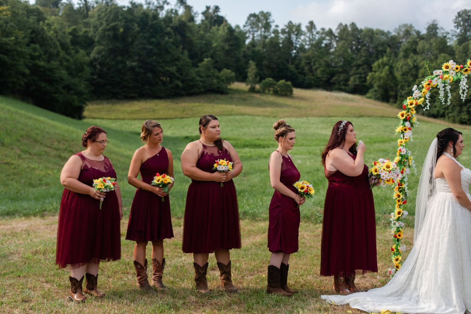 Wedding party in burgundy dresses and casual attire poses in line on grassy hillside.
