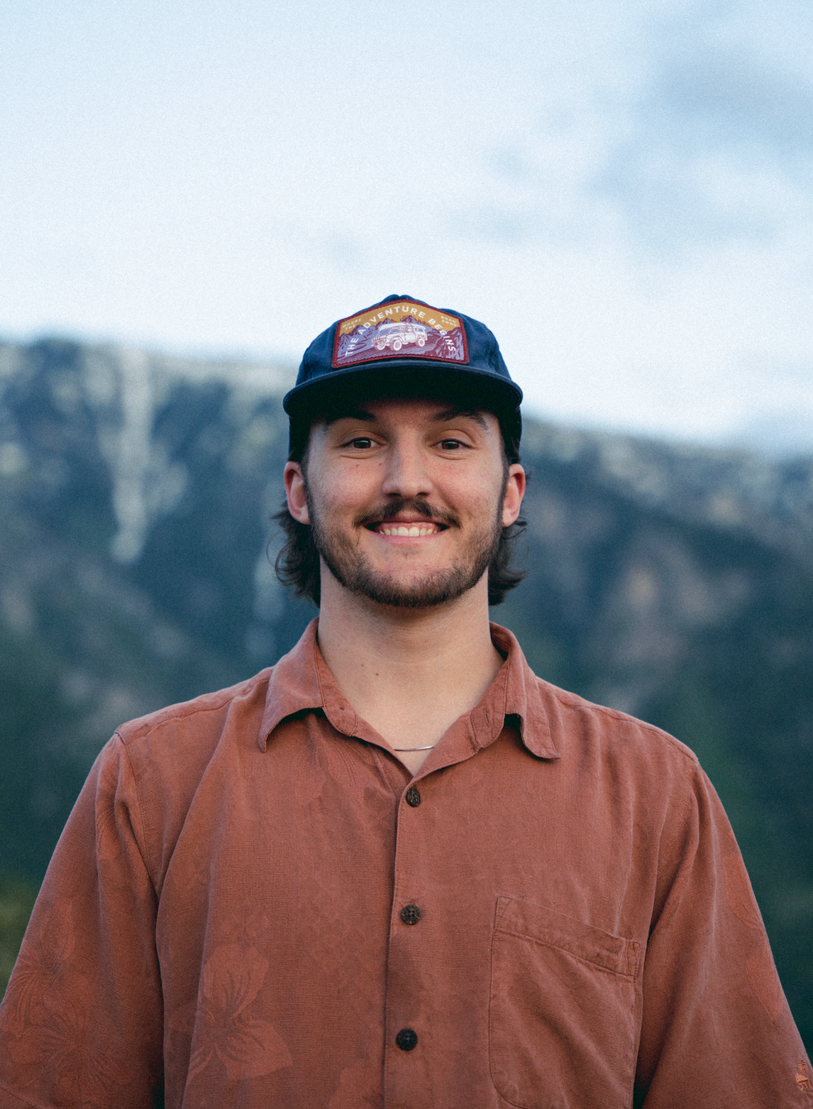 A person in a baseball cap smiles at the camera with mountain peaks in the background.