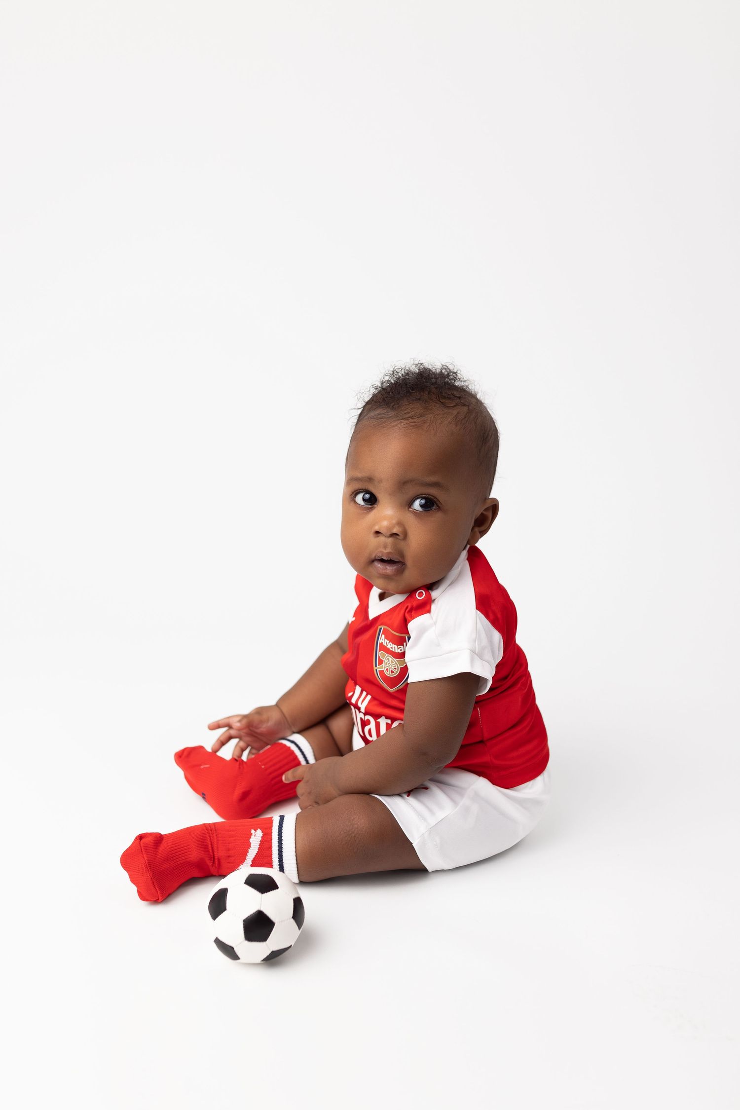 Baby in red and white outfit sitting with soccer ball against white background.