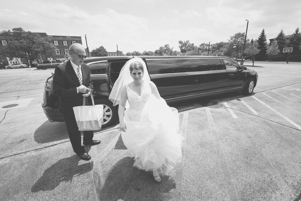 Black and white photo of a bride entering a vintage limousine on wedding day.