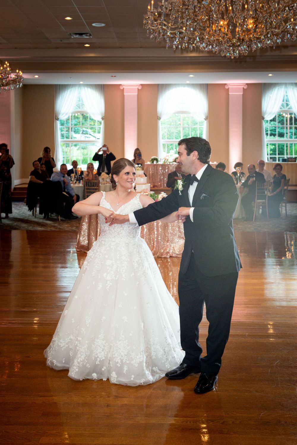 Couple sharing their first dance on a polished wooden dance floor under crystal chandeliers.