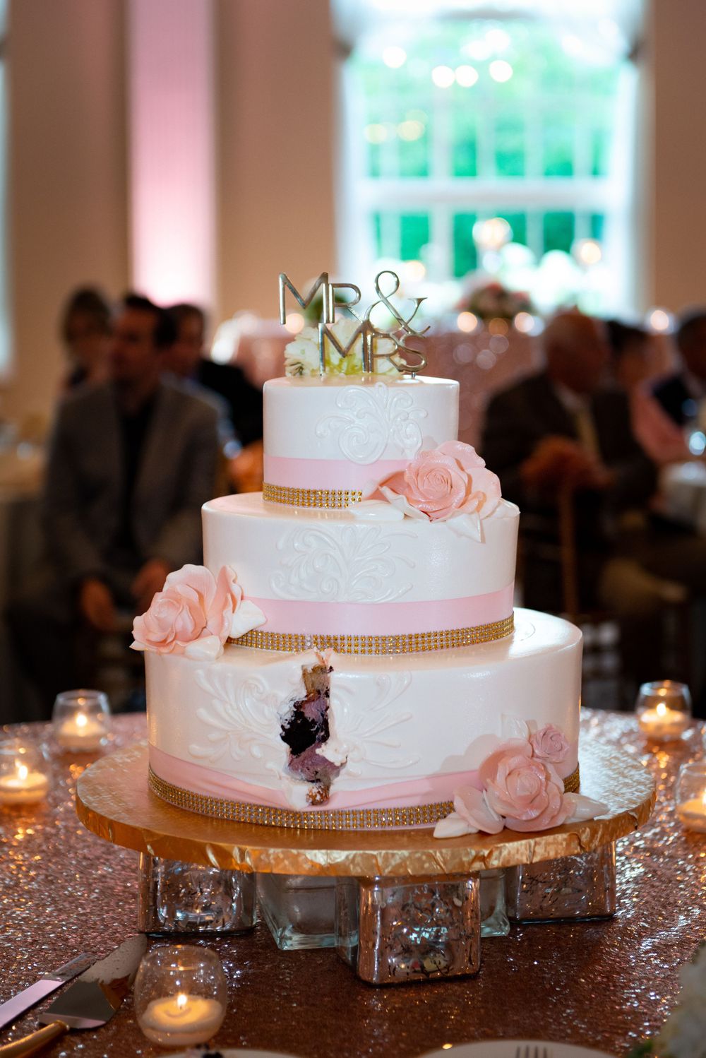 Three-tiered white wedding cake decorated with blush pink roses and Mr & Mrs toppers on a sparkly gold stand.