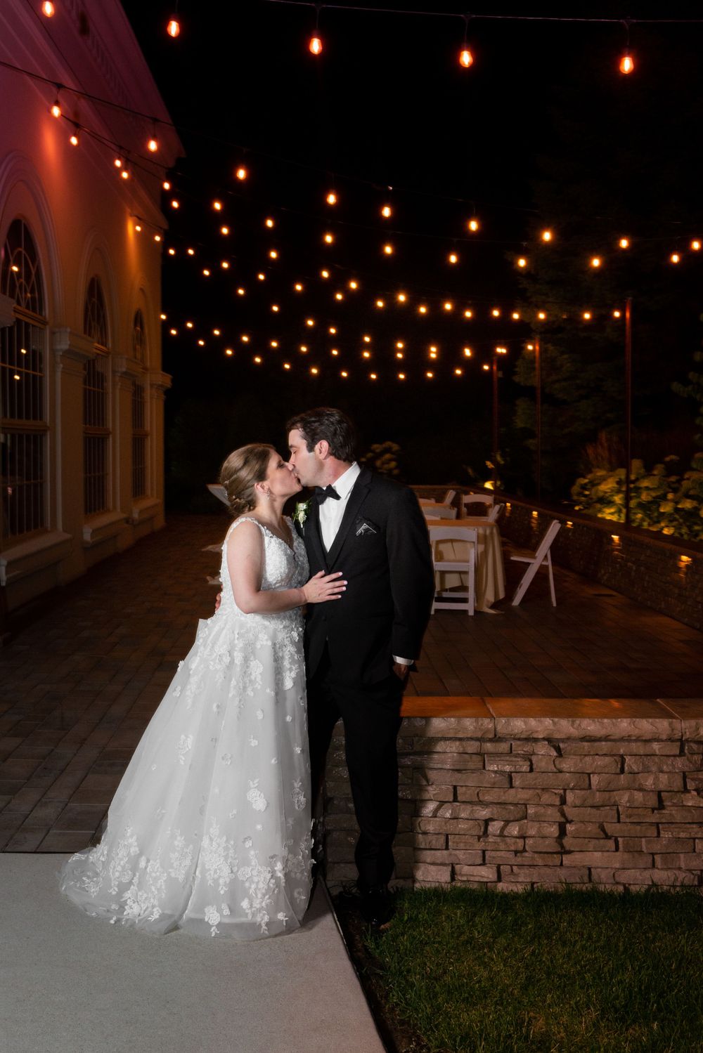 Couple sharing an intimate moment under twinkling string lights on a stone patio at night.
