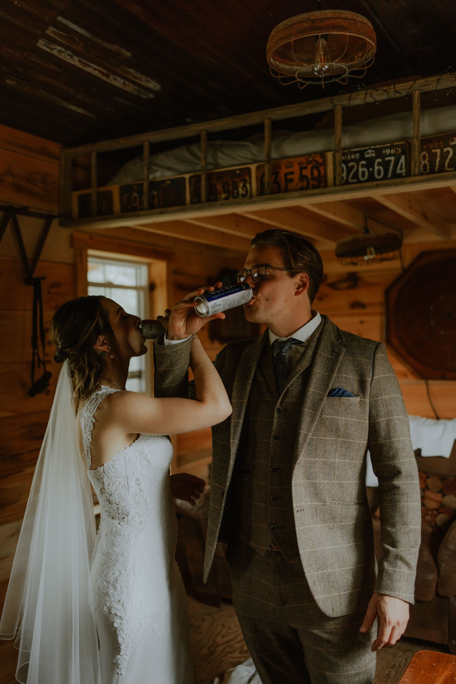 a bride and groom linking arms while drinking beer in a cabin