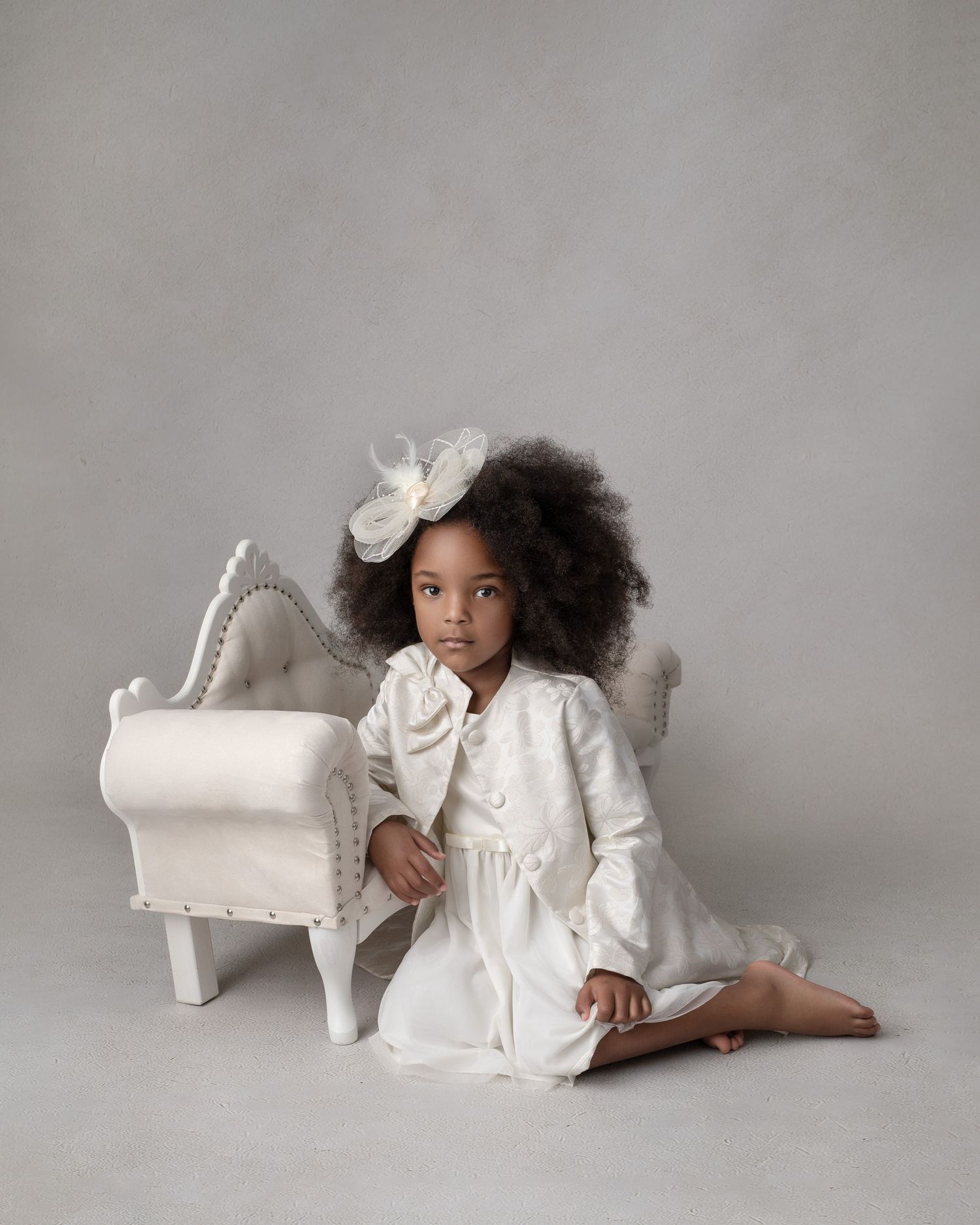 Child in white dress posing elegantly beside miniature chair against neutral gray backdrop.