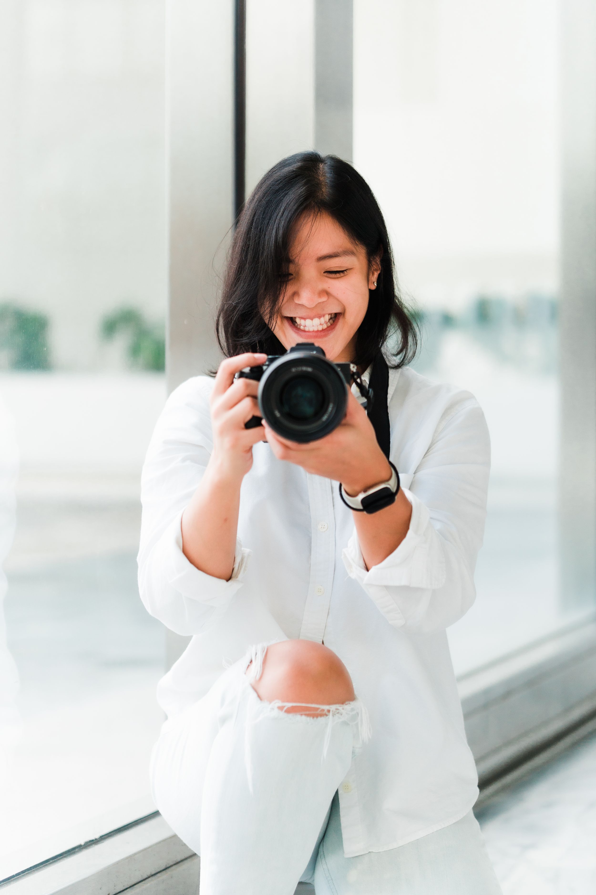 Professional photographer in white outfit smiling while holding camera in bright indoor setting.