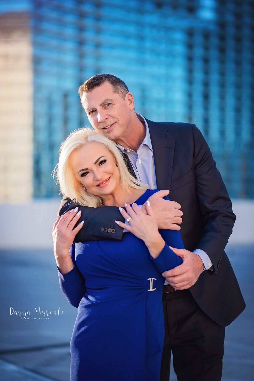 Couple in formal attire embracing against modern blue building background.