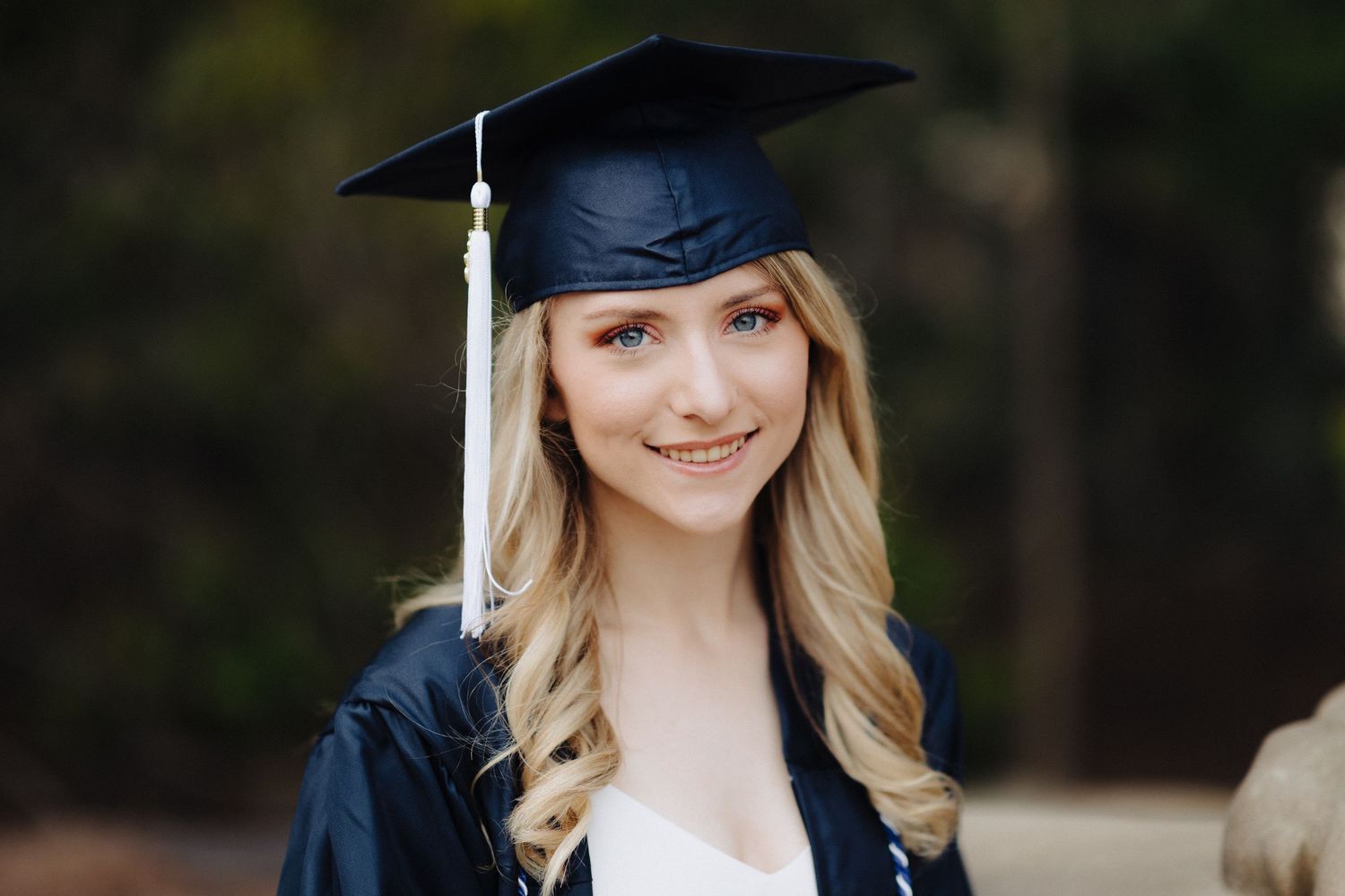 Graduation portrait of person in cap and gown with white tassel against blurred background.