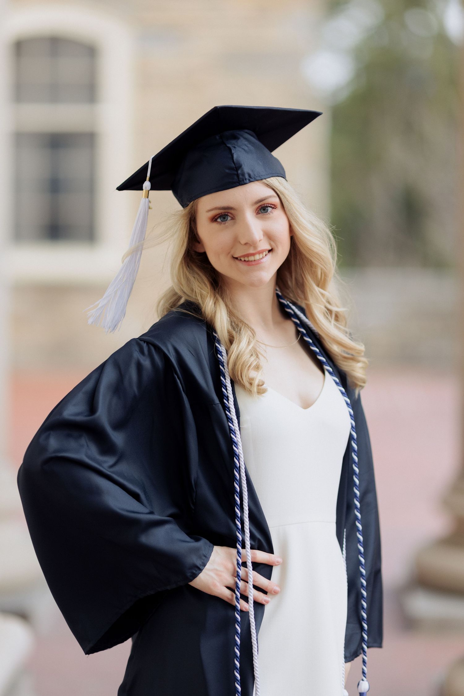 Graduate in traditional cap and gown with white dress poses for portrait outdoors.