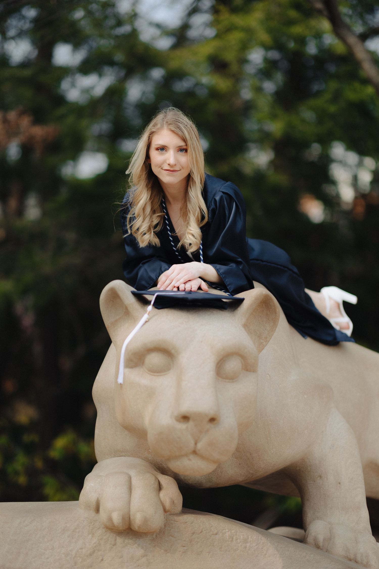 Graduate in academic regalia sits on iconic Penn State Nittany Lion statue during photo session.