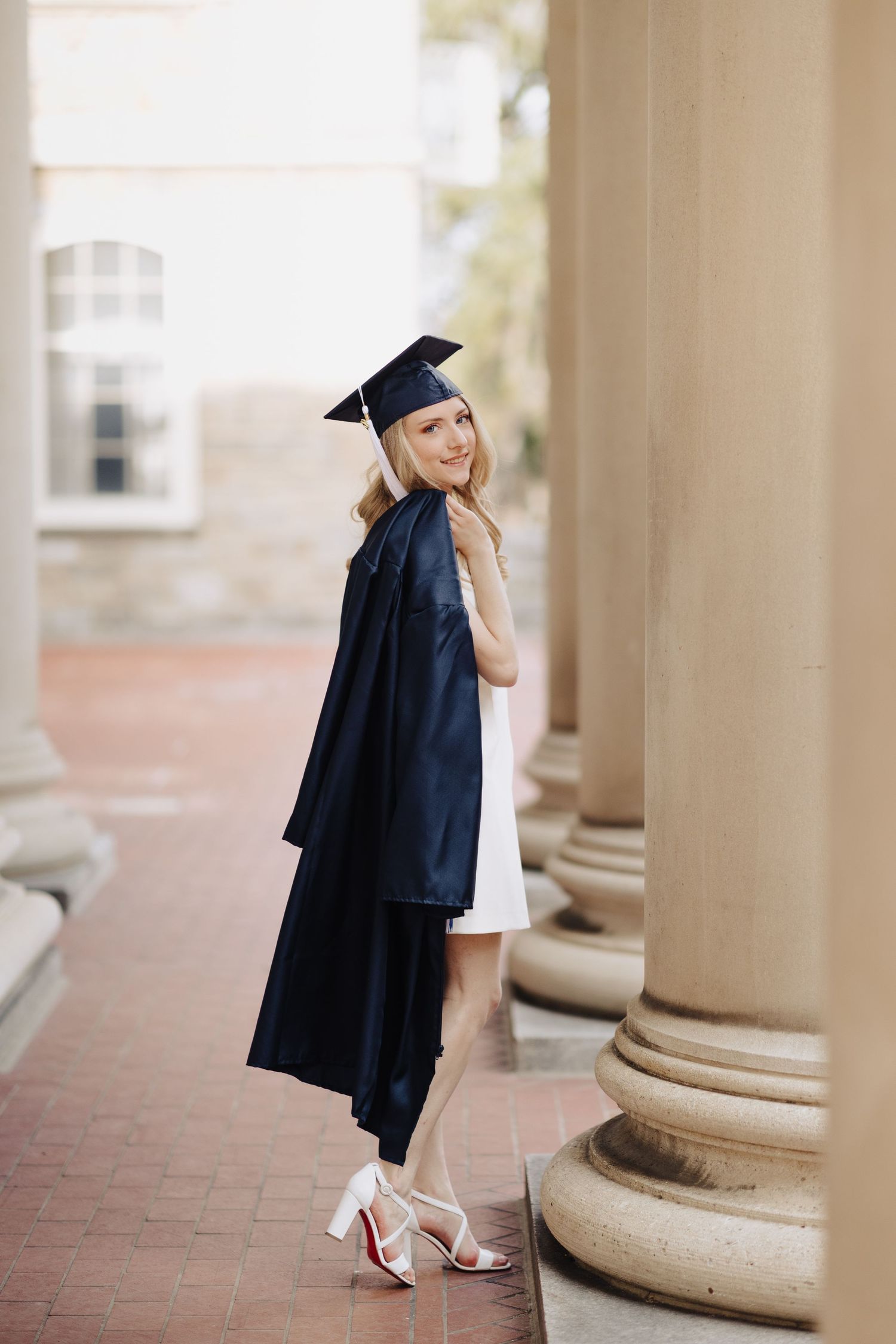 Graduate in academic regalia stands between pillars on brick walkway glancing over shoulder.