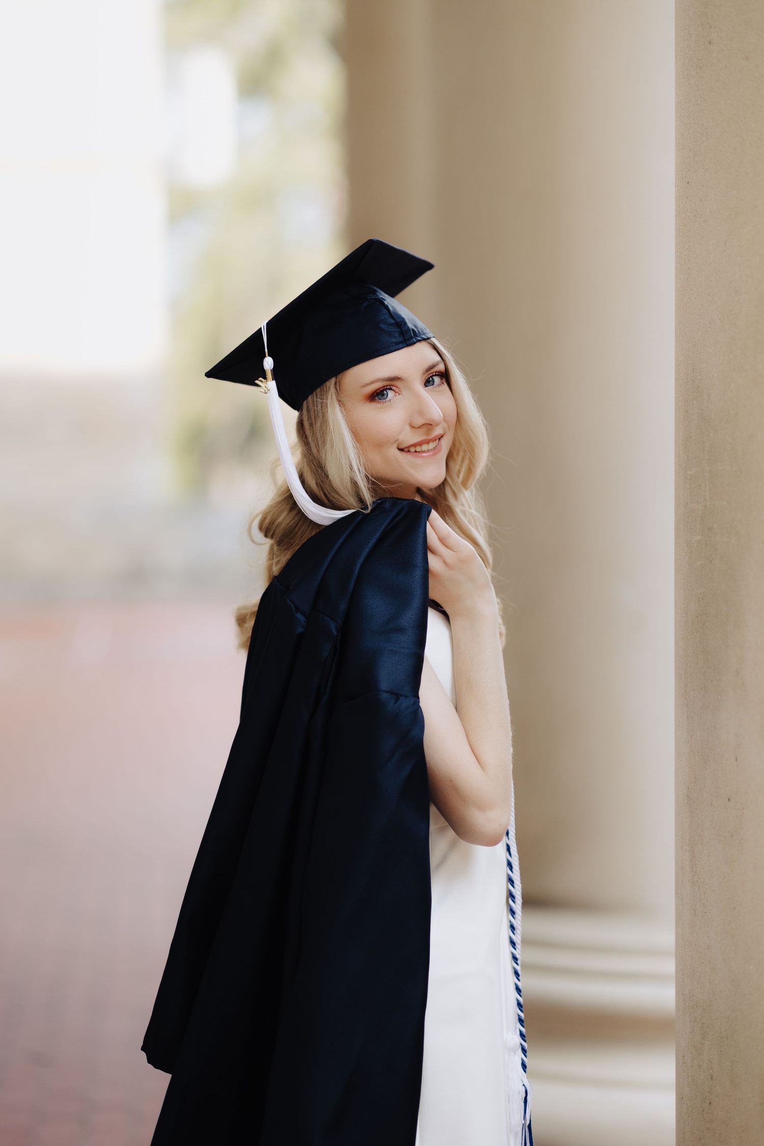 Graduate wearing academic regalia smiles next to stone columns during graduation photoshoot.