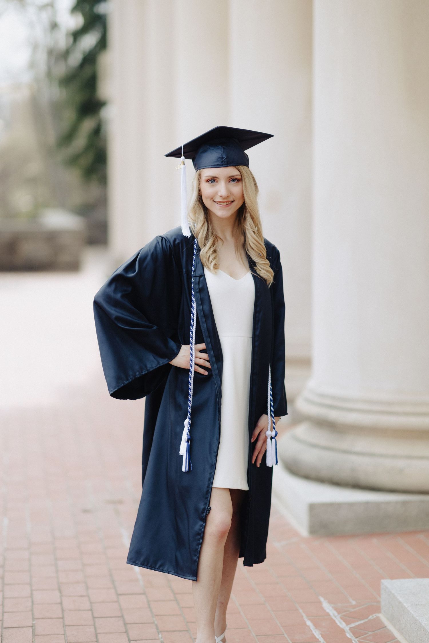 Graduate in navy blue cap and gown poses outdoors by classical columns in white dress.