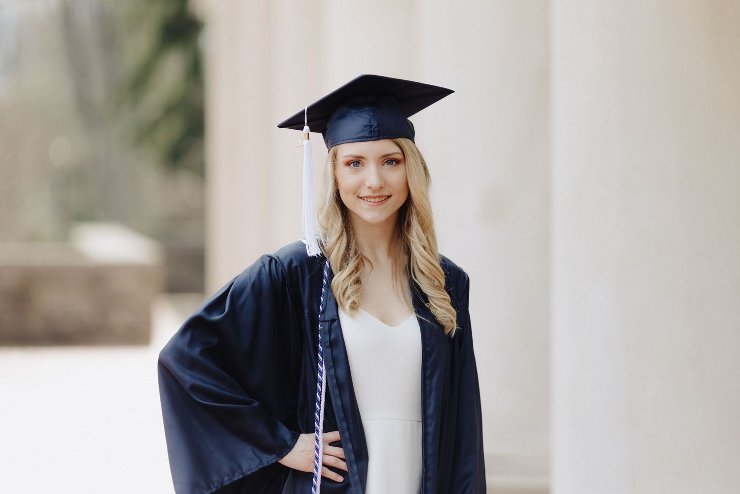 Graduate in traditional navy cap and gown poses against white columns.
