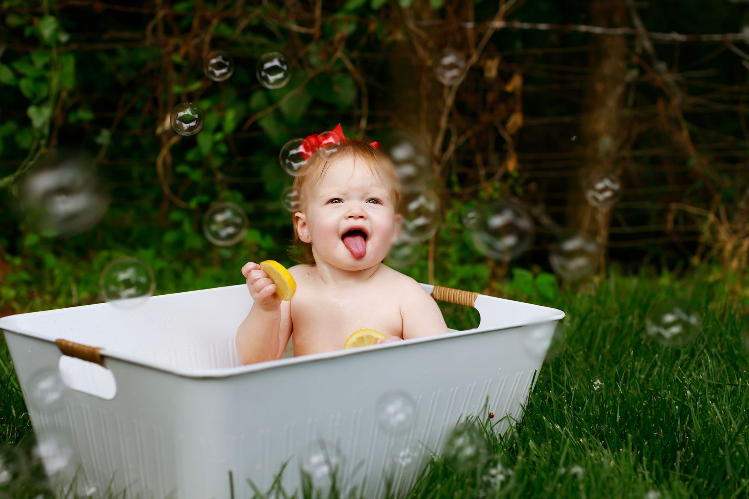 Young child plays in white tub outdoors while soap bubbles float in garden setting.