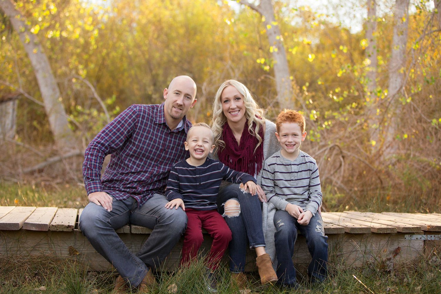 Happy family of four sits together on wooden bench during autumn sunset.