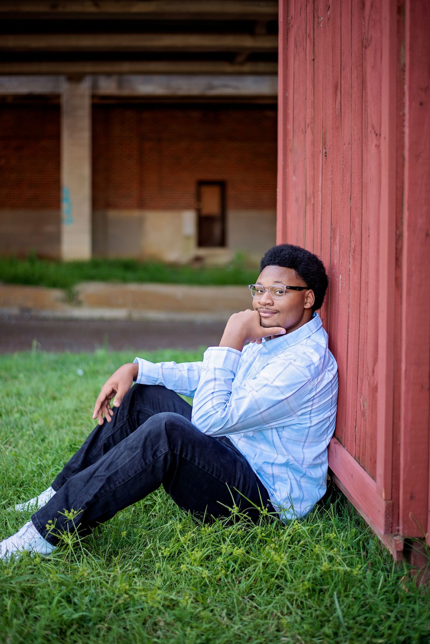 Student in light blue shirt and glasses poses casually against red barn wall.