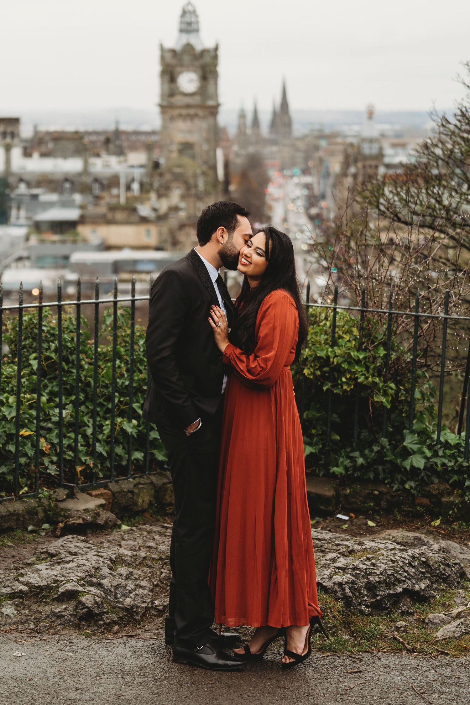 Romantic couple embracing against Edinburgh cityscape with historic architecture in background.