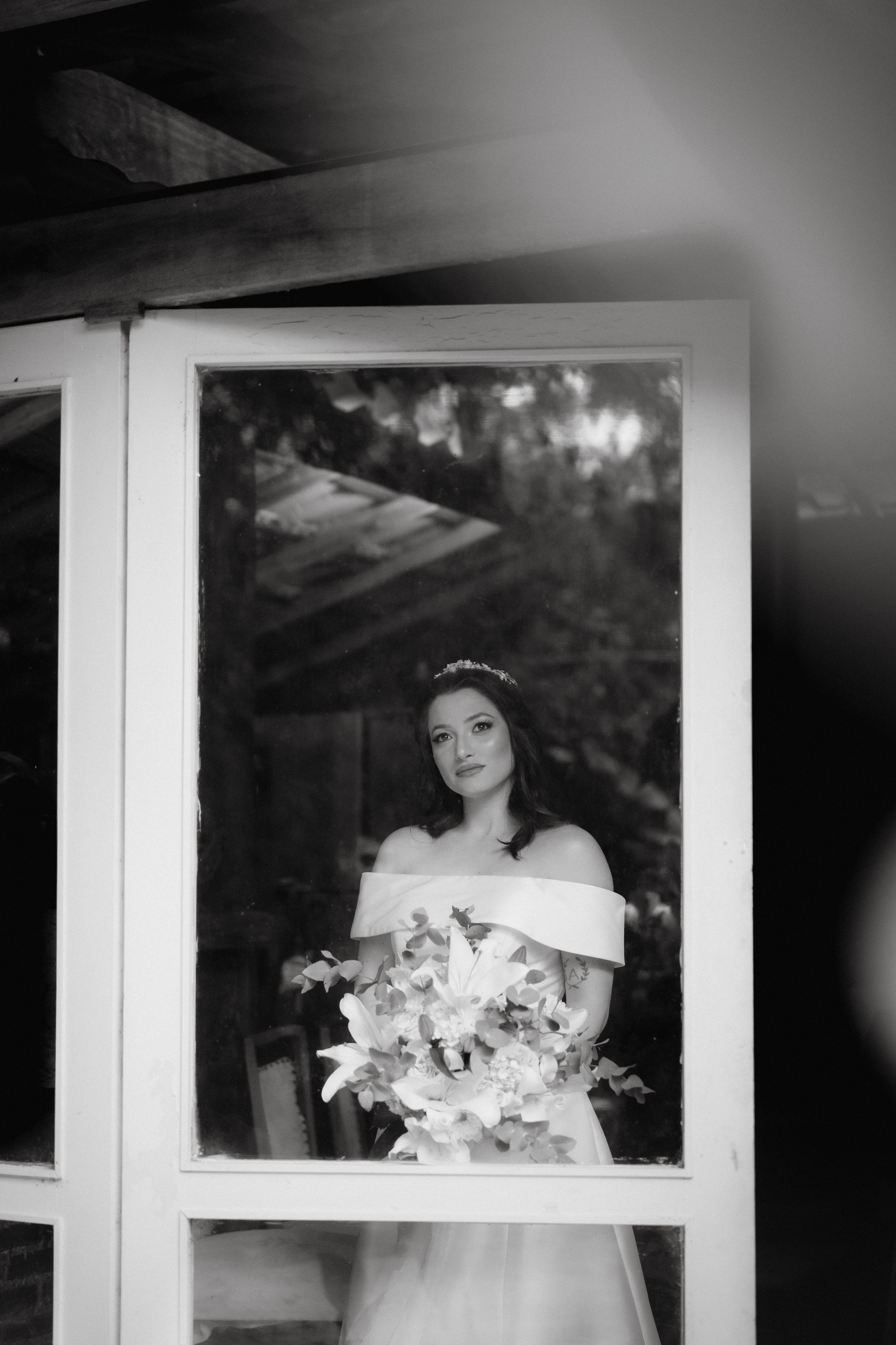 A bride in an off-shoulder white gown holds a bouquet while standing behind a window frame in black and white.